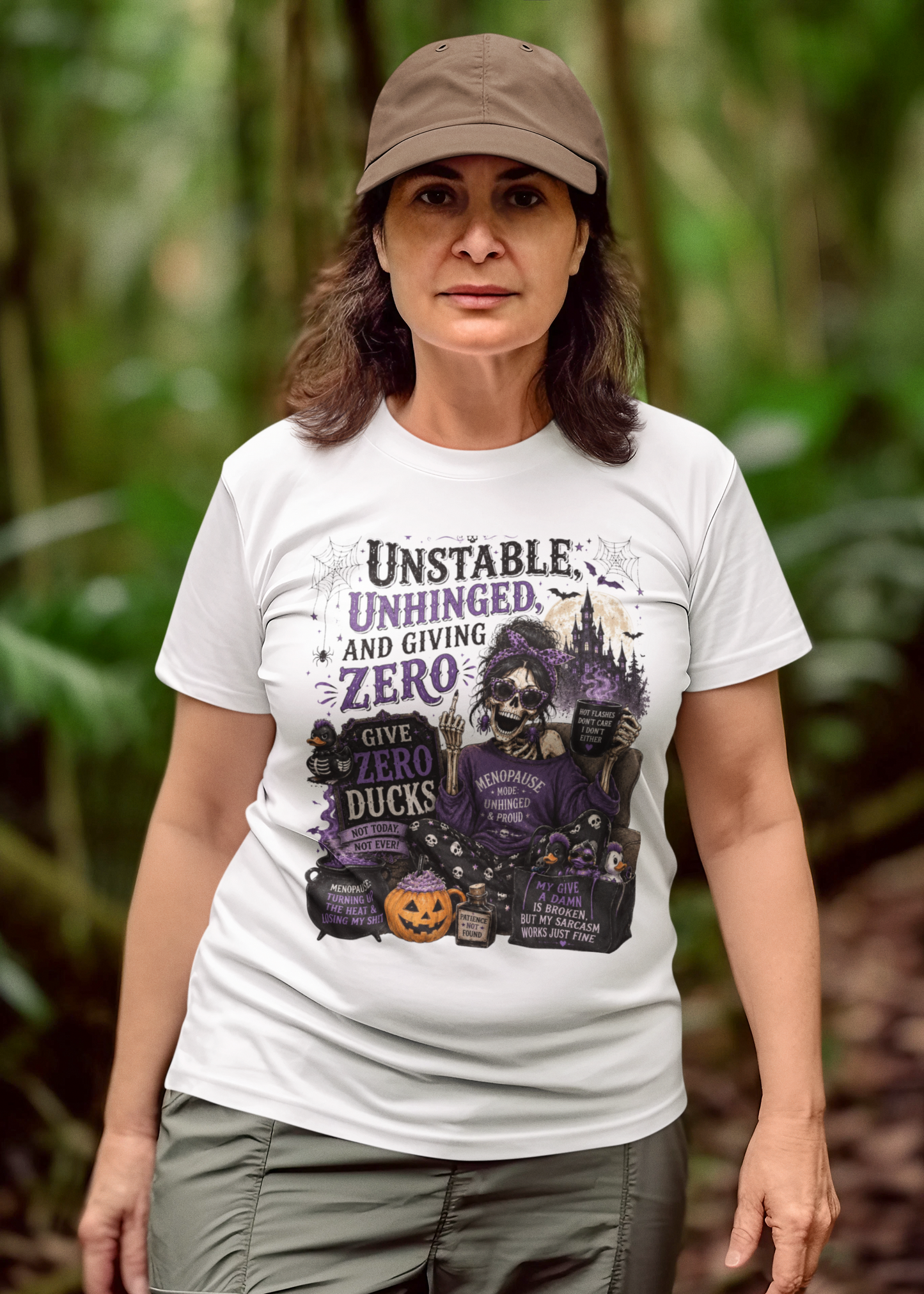 A woman wearing a white graphic t-shirt with a Halloween-themed design, standing in a lush green forest. The graphic features a witch, pumpkins, and the text 'Unstable, Unhinged, and Giving Zero'.