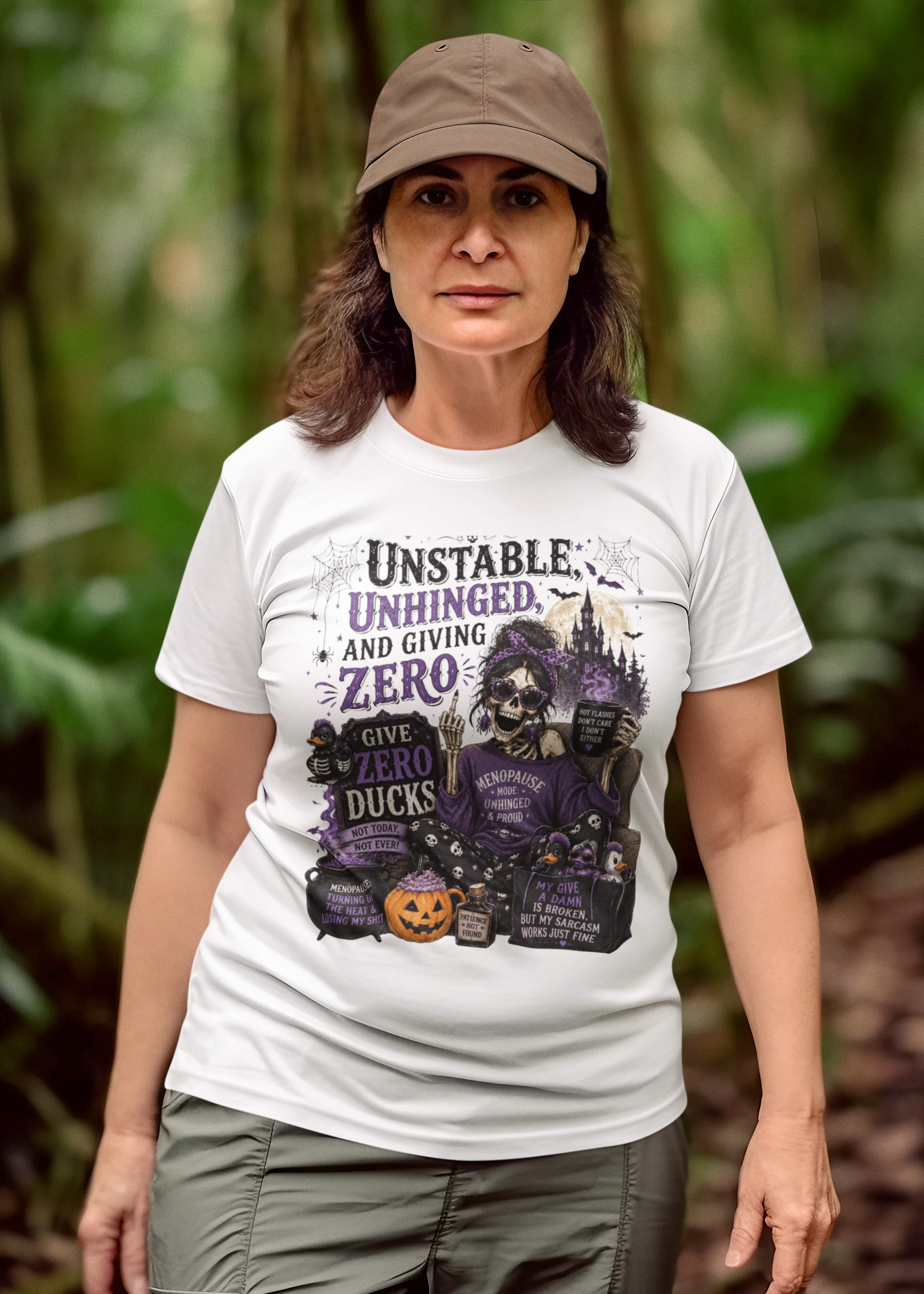 A woman wearing a white graphic t-shirt with a Halloween-themed design, standing in a lush green forest. The graphic features a witch, pumpkins, and the text 'Unstable, Unhinged, and Giving Zero'.
