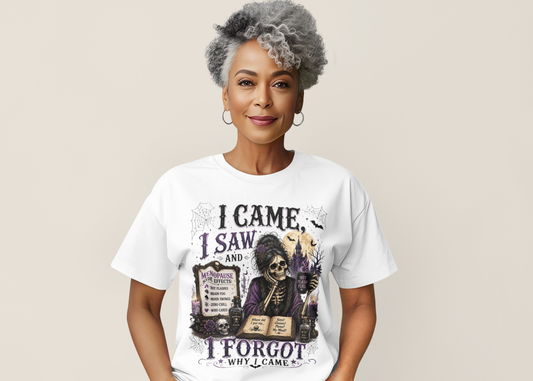 A woman with styled gray hair wearing a white graphic t-shirt with a Halloween-themed illustration of a skeleton and the text 'I CAME, I SAW, AND I FORGOT WHY I CAME'.