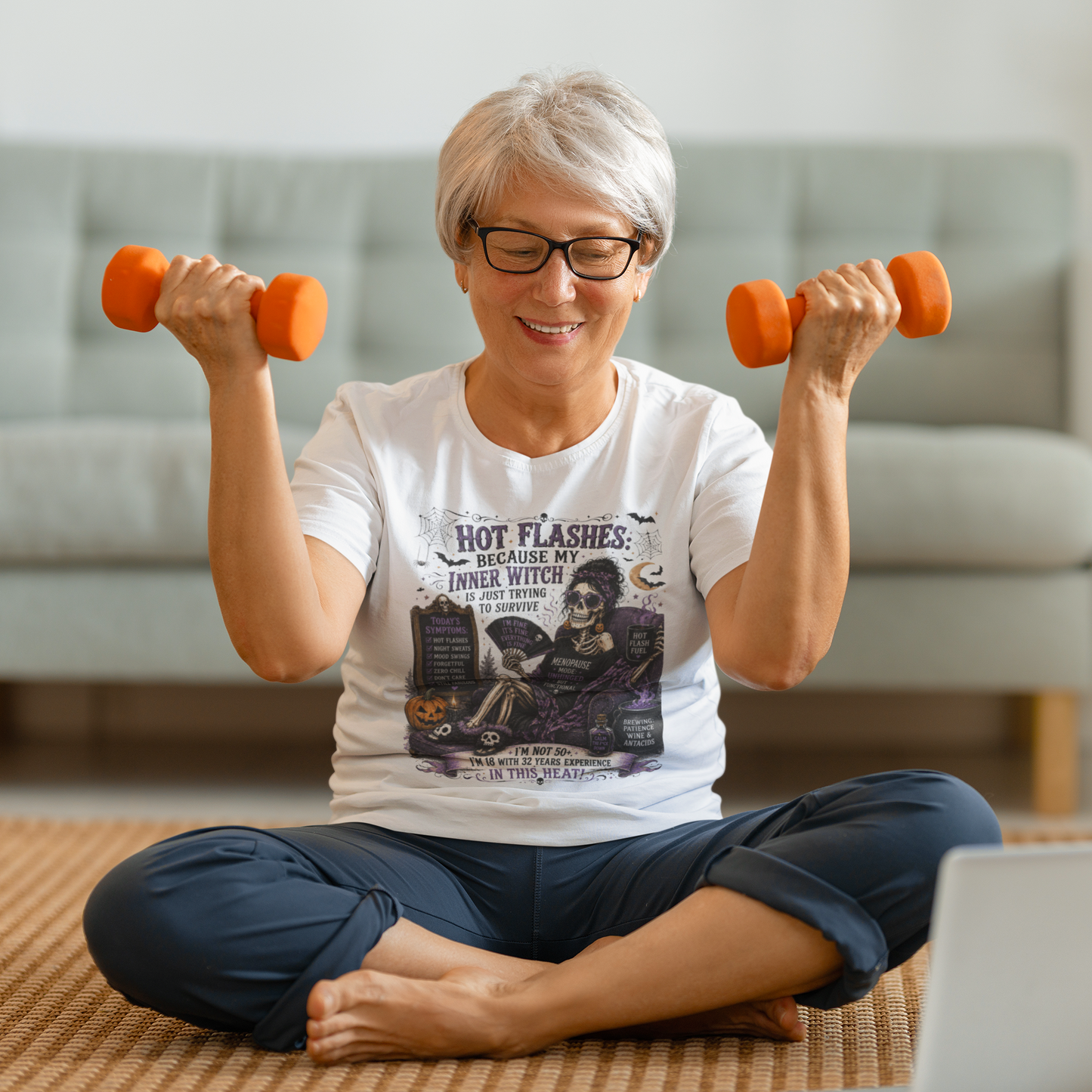 A woman with short gray hair and glasses sits cross-legged on a woven rug, lifting orange dumbbells. She is wearing a white graphic t-shirt with a gothic skeleton design and dark navy pants in a brightly lit living room with a gray sofa in the background.