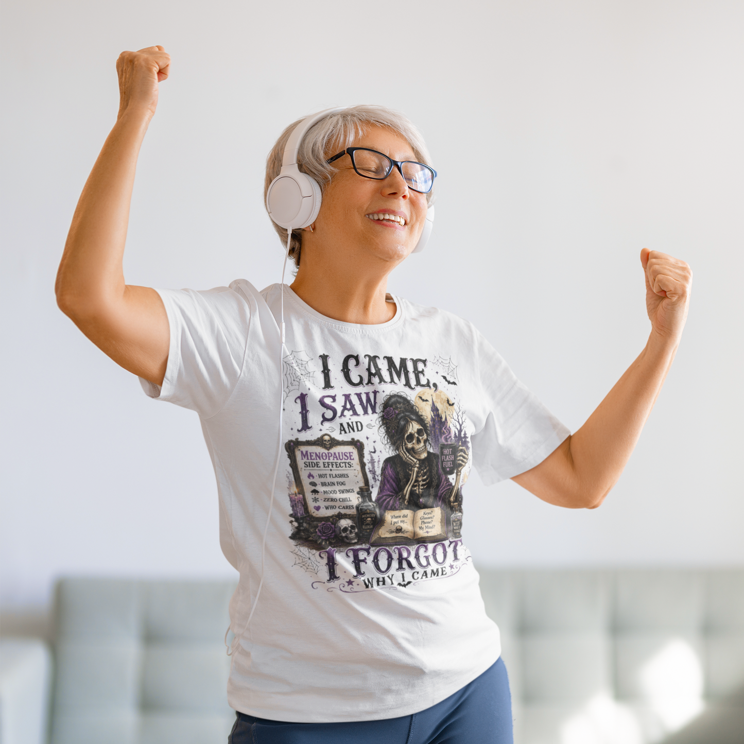 An older woman with short gray hair, glasses, and white headphones is celebrating with raised fists, wearing a white graphic t-shirt with a skeleton illustration and the text 'I CAME, I SAW, AND I FORGOT WHY I CAME'.