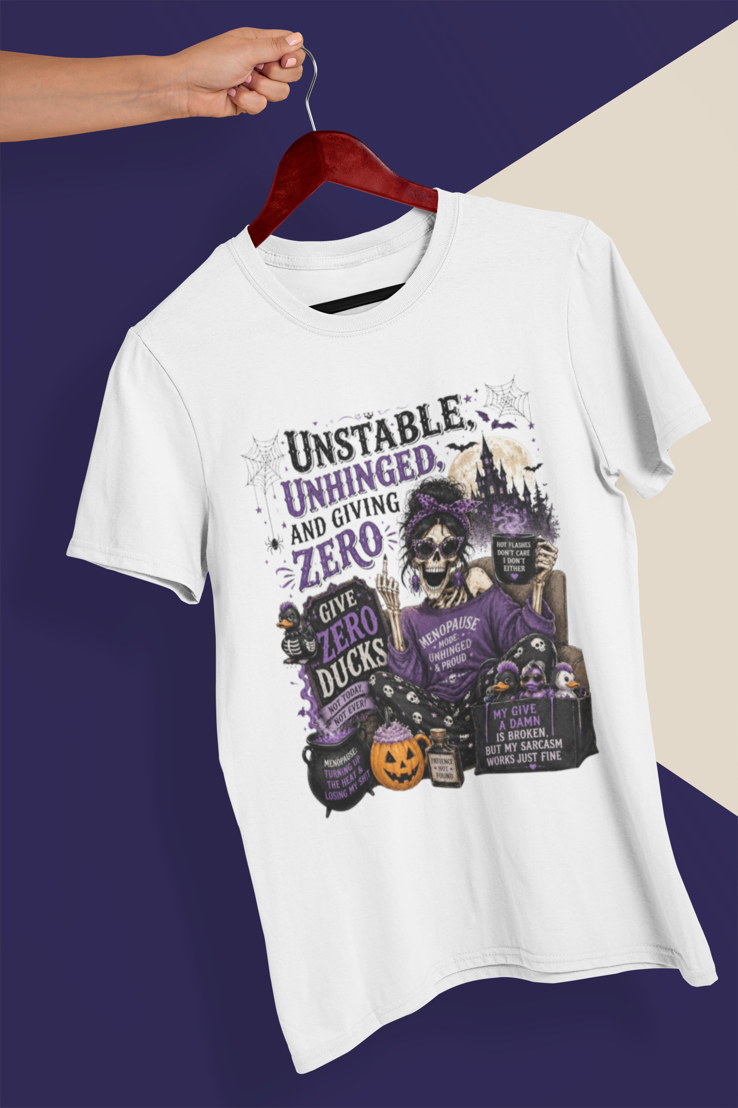A white short-sleeve graphic T-shirt hanging on a dark red wooden hanger held by a person's hand, against a split dark purple and beige background. The shirt features a Halloween-themed graphic with the text 'Unstable, Unhinged, and Giving Zero' and a skeleton character in purple clothing surrounded by pumpkins, cauldrons, and tombstones.