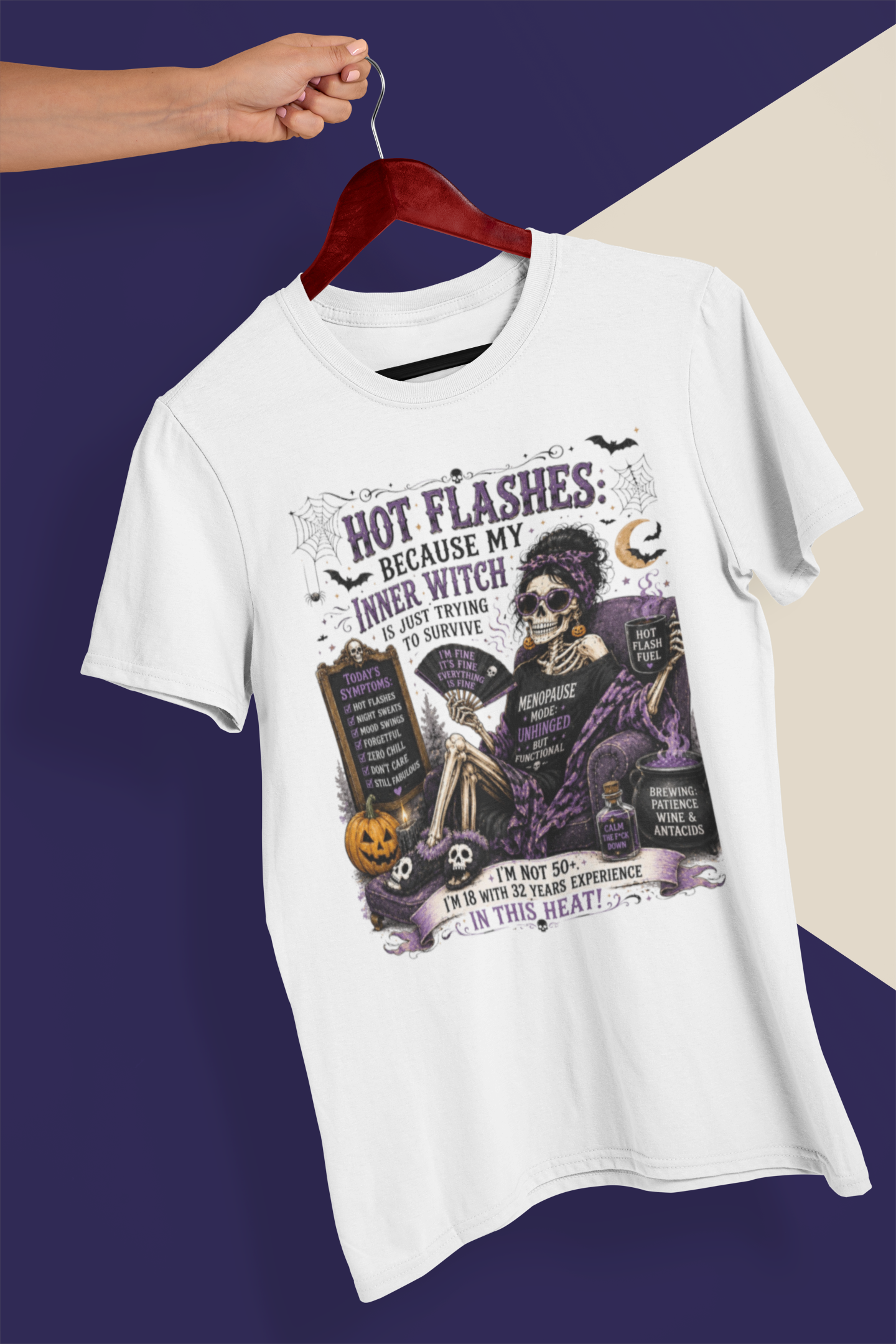 A white short-sleeve graphic T-shirt hanging on a dark red wooden hanger, held by a person's hand against a split dark purple and beige background. The shirt features a detailed Halloween-themed graphic with a skeleton, tombstones, pumpkins, and gothic text.
